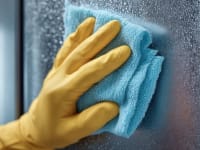 A person wearing gloves wipes a stainless-steel fridge with a microfiber towel.