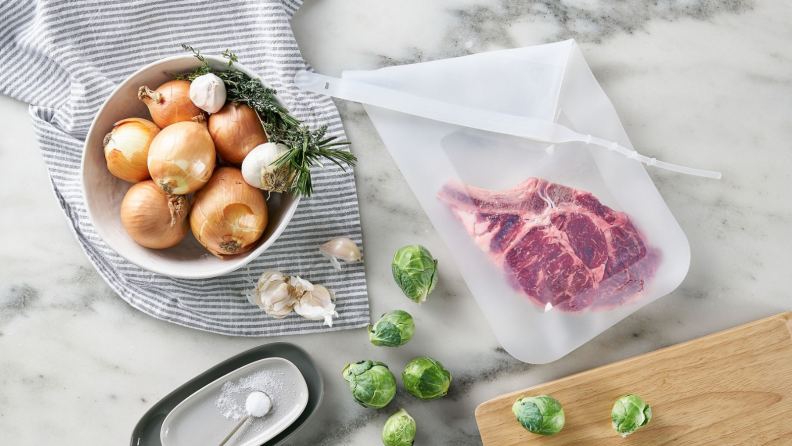 A piece of steak is inside a Stasher silicone bag surrounded by a bowl of onions, garlic, herbs, Brussels sprouts.