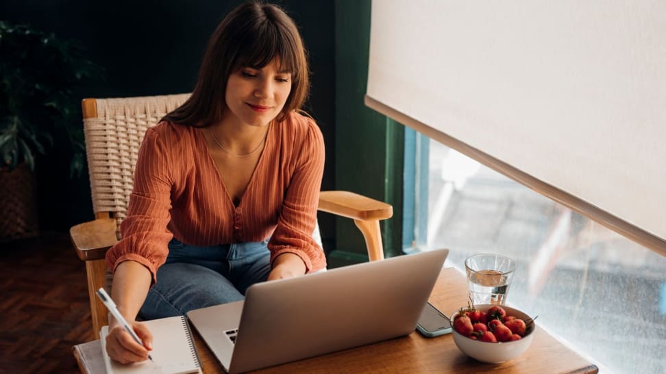 Person writing in notebook while sitting in front of a laptop on a desk near glass of water and bowl of fruit