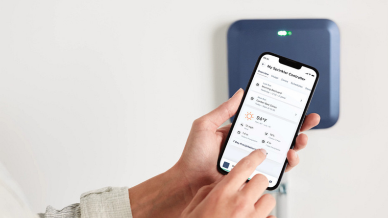 A person holding a phone showing the Moen Smart Water app in front of the Moen Smart Sprinkler Controller.