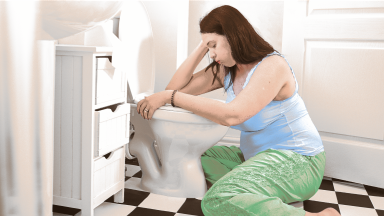 A pregnant person rests over a toilet, sick with morning sickness.