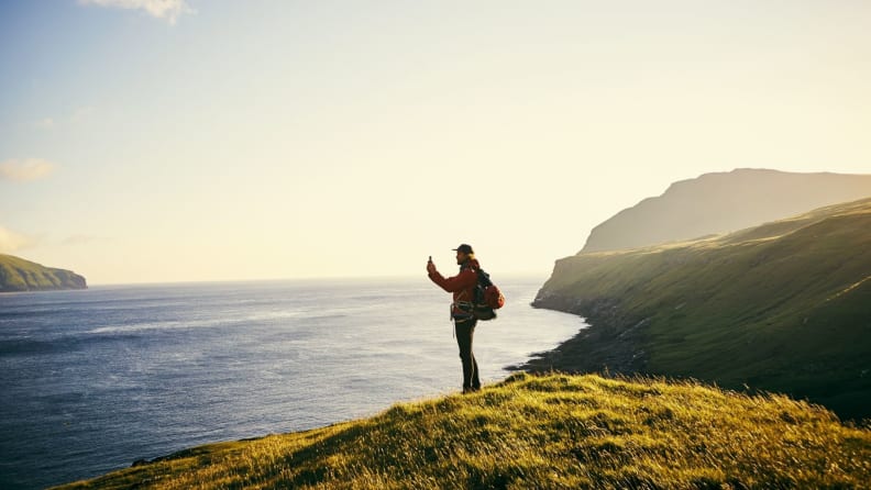 A hiker with a backpack stands on a cliffside to snap a photo of the sea