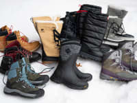 A number of pairs of winter boots, from various brands, laid out on a bed of snow.