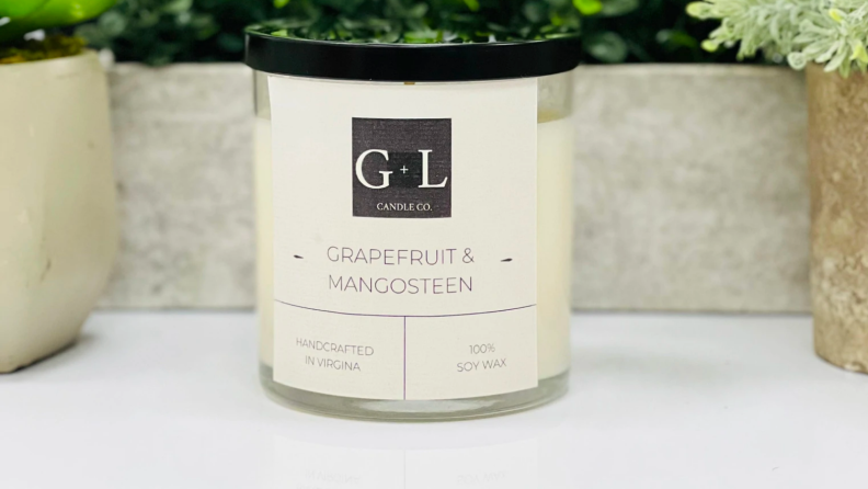 An image of a "Grapefruit and Mangosteen" candle in a clear jar with a black top, surrounded by plants.