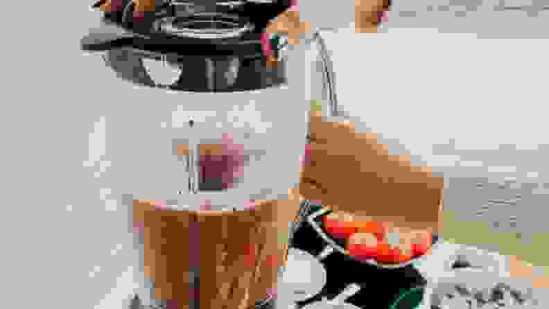 A woman with painted nails uses a blender to make soup. Tomatoes and basil sit on the countertop next to the blender.