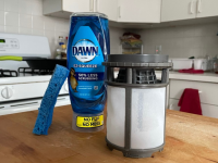 A dishwasher filter next to a bottle of dish soap and a sponge.
