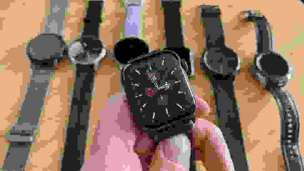 A hand holding an Apple Watch. In the background are several smartwatches laid out on a table.