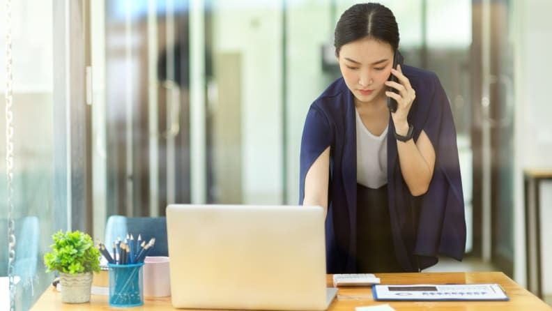 Woman dressed in professional attire in front of laptop, using phone and working.