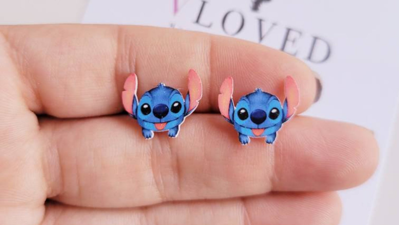 A hand holding two blue Stitch earrings