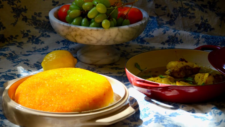 A plate of golden tahdig in the noosh-e-joon platter is in the center of the image. In the shadow, there's a plate of fruits and a pot of saffron chicken.