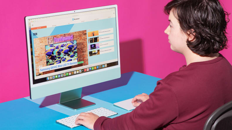 A person using an all-in-one iMac desktop computer that stands on a blue surface against a hot pink background