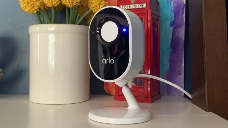 The white, oval-shaped Arlo Indoor Wireless Camera sits on a white desk shelf.