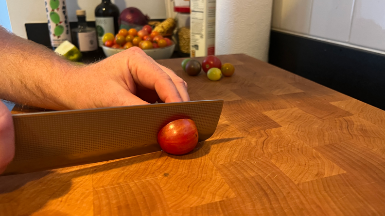 A person using the Milk Street Nakiri knife slicing a cherry tomato in half on top of a wooden cutting board.