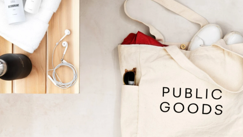 Filled canvas tote bag with "Public Goods" written on front, next to small wooden table.