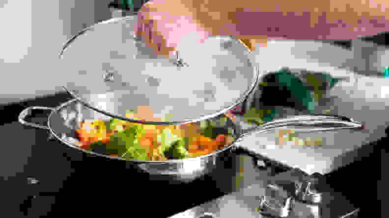 A person is lifting the lid off of a wok full of cooked vegetables.