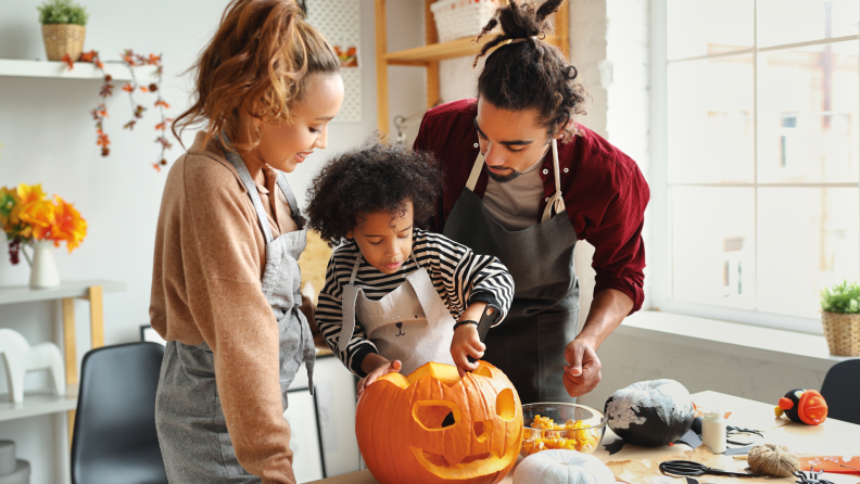 Two parents supervise small child in kitchen as they carve a pumpkin together.
