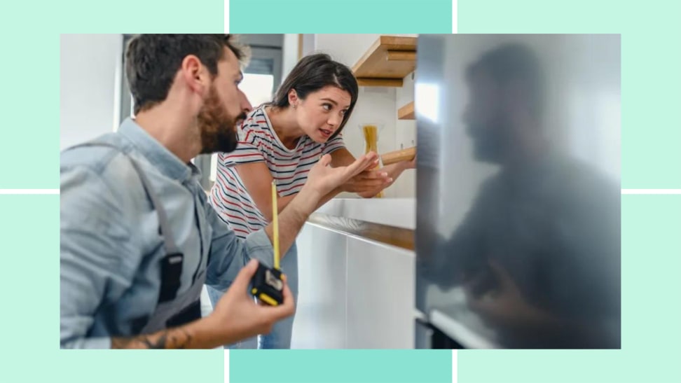 A man and woman stand next to a refrigerator. The man has a tape measurer and the woman is gesturing towards the fridge, which sticks out beyond the cabinetry.