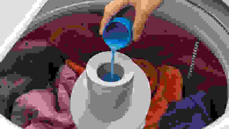 A shot of the interior drum, full of colorful laundry, with pole agitator sticking out of the center. A hand pours fabric softener into the center of the pole agitator.