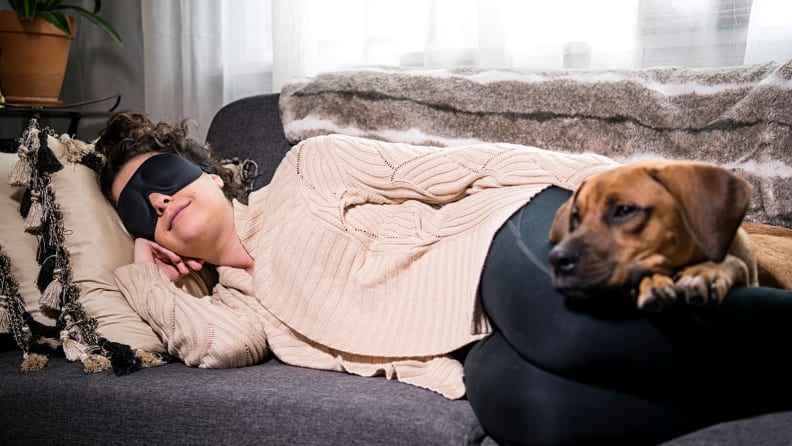 A person laying on the couch wearing a sleep mask with their dog.