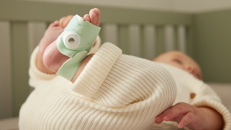 A small infant laying on their back in their crib while wearing a mint green Dream Sock on one foot.