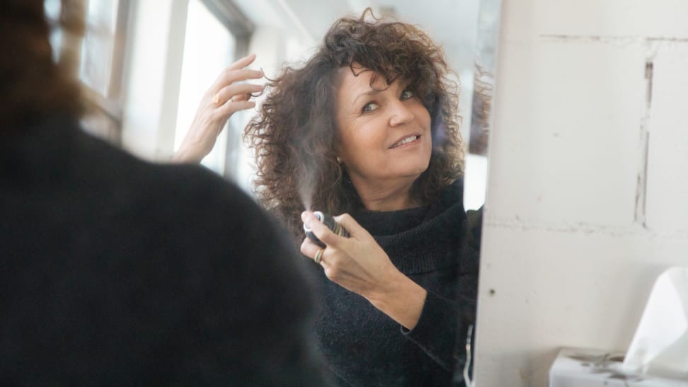 A woman spraying hairspray while looking in a mirror