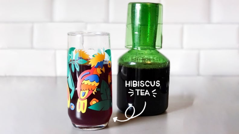 A colorful glass filled with iced hibiscus tea next to a green glass carafe filled with more of the same.