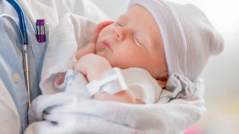 A Caucasian female infant is asleep in the arms of a pediatrician. The baby is swaddled in a blanket and is wearing a warm cap.