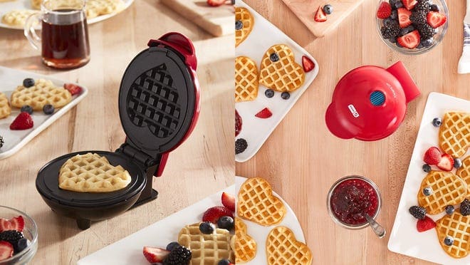 Small, red waffle maker surrounded by heart-shaped waddle with assorted fruits in bowl.