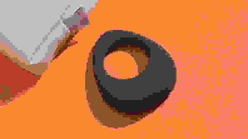 A black Maude Vibe vibrating cock ring next to a tan pouch on top of an orange surface.