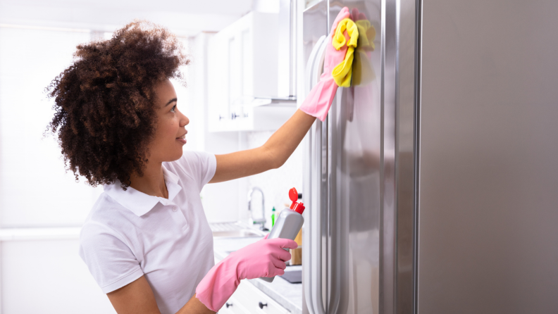 Person wearing rubber gloves wipes the outside of a refrigerator with a cloth.