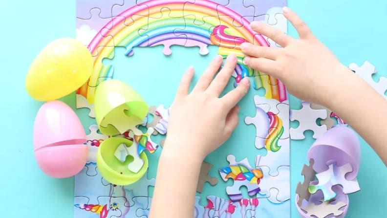 A child's hands piece together a rainbow printed puzzle alongside opened Easter eggs.
