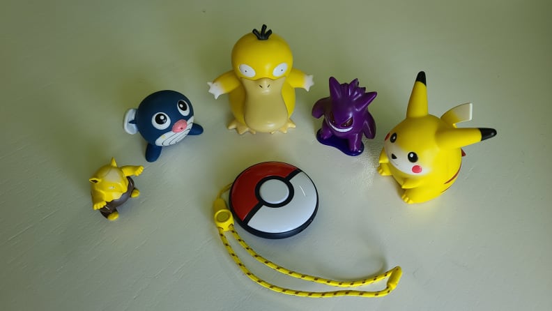 Pokemon toys and keychain in a circle on a white counter, surrounding the Pokémon Go Plus +.
