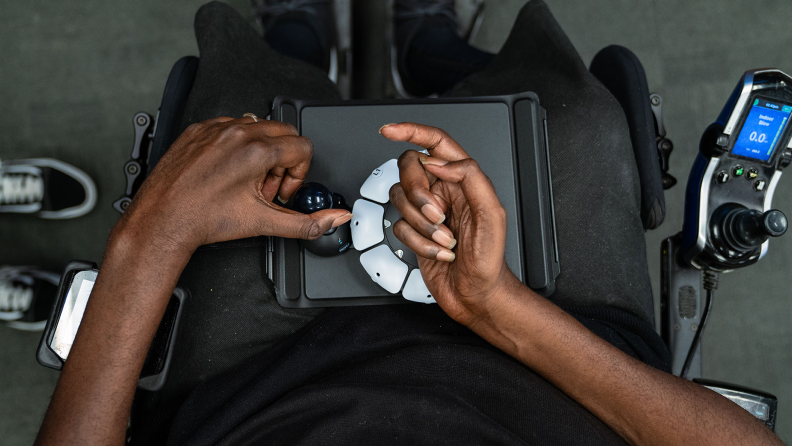 Person with limited mobility using hand to operate the buttons on the PS5 Access Controller whiles sitting in wheelchair.