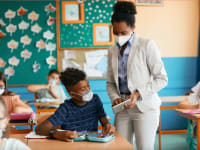 Teacher wearing face mask while helping student in face mask in classroom setting.