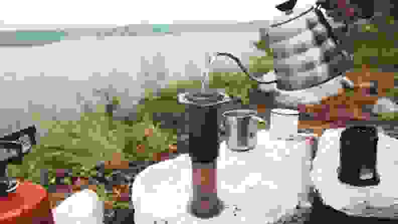In what appears to be a riverbank, a person is pouring water into an Aeropress coffee maker. A portable stove is next to the Aeropress.