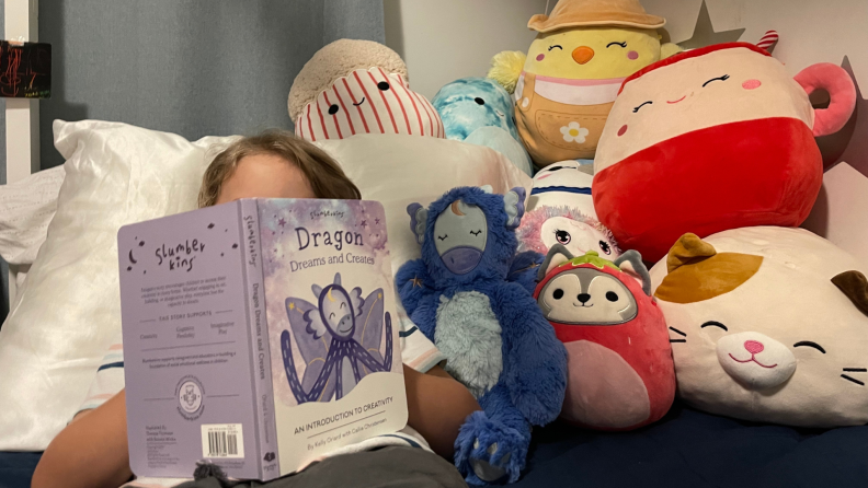 Small child laying down in bed against pillow next to Slumberkins Dragon Kin and other stuffed animals while holding book in front of face.