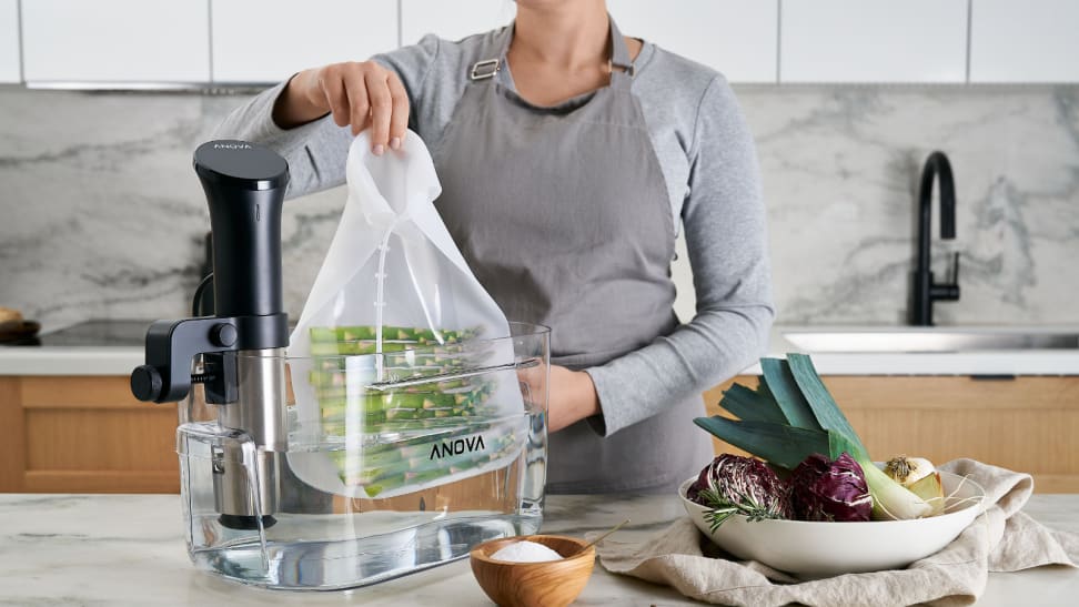 A woman is putting a bag of asparagus into a half-filled water tank for sous vide. An immersion circulator is attached to the tank to control the temperature of the water.