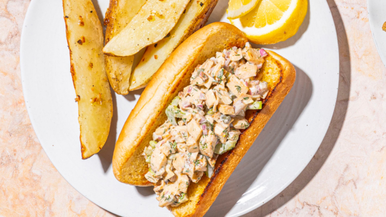 An image of a "lobster" roll made with heart of palm, on a plate with fries and lemon wedges.