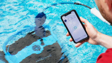 Robotic pool vacuum in a swimming pool with a person holding a smartphone showing the app in the foreground.