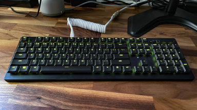 A Razer Deathstalker V2 Pro keyboard lit with green accent lights sits on a wood desk.
