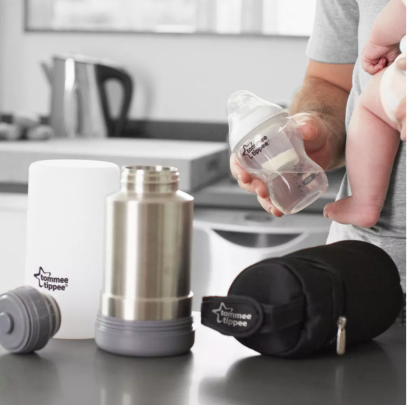 The Tommee Tippee travel bottle warmer system is displayed on a counter, in the background a parent holds a baby on a hip and an empty bottle in their other hand.