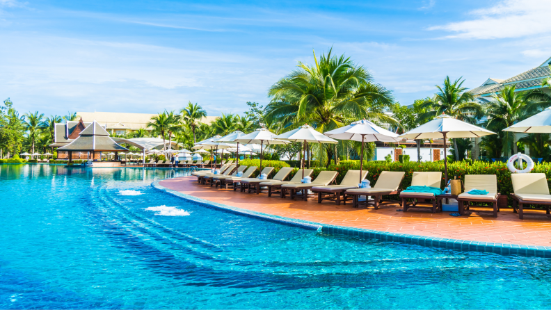 A luxurious resort pool with lounge chairs, umbrellas, and palm trees under a bright blue sky.