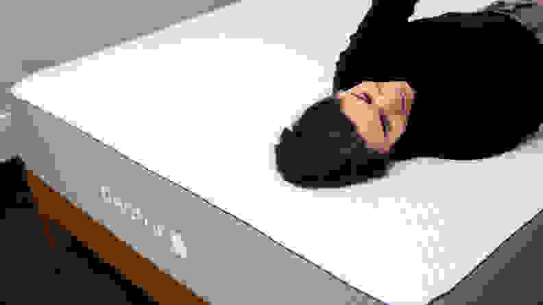 A person laying down on a bare quilted mattress that sits on a wood bed frame in a carpeted room
