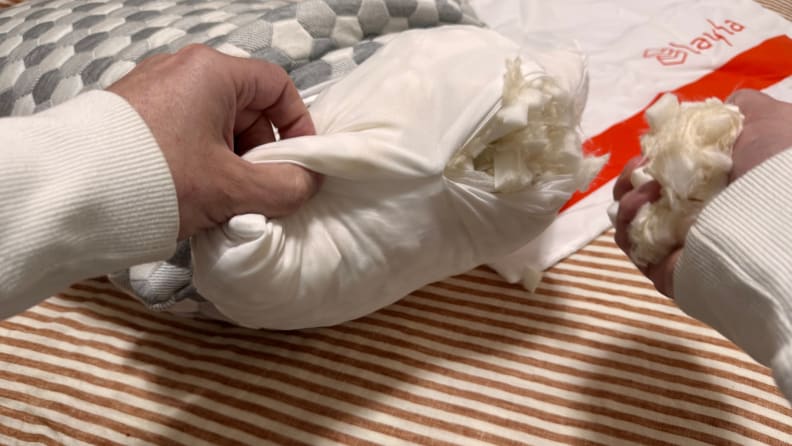 Layla Sleep Kapok Pillow being held open to see the inside filling, in front of a striped white and brown background.