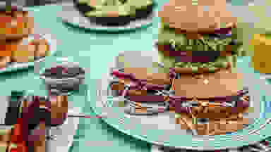 A spread with vegan burgers, skewers, and other out-of-focus plates with food on a blue tablecloth