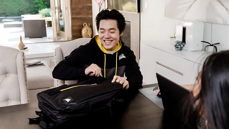 A young man smiles while unzipping a Corsair backpack at a table during a casual work-from-home setup.