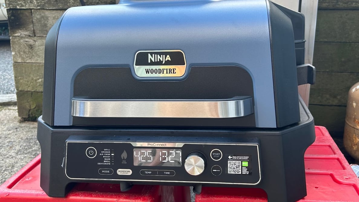The Ninja Woodfire ProConnect XL Outdoor Grill & Smoker in black and stainless steel finish sits on top of red surface outdoors with lid closed.