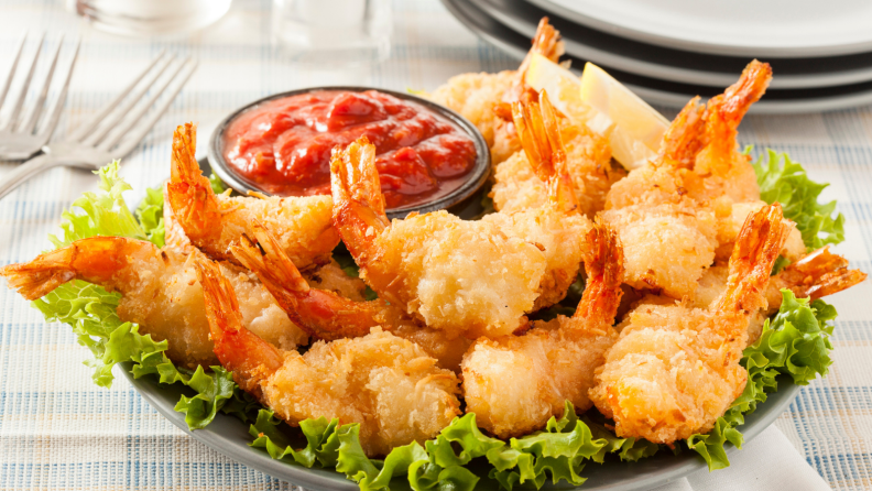 A plate of fried coconut shrimp and a side of sauce on a bed of lettuce, surrounded by a set table, with forks and plates in the background.