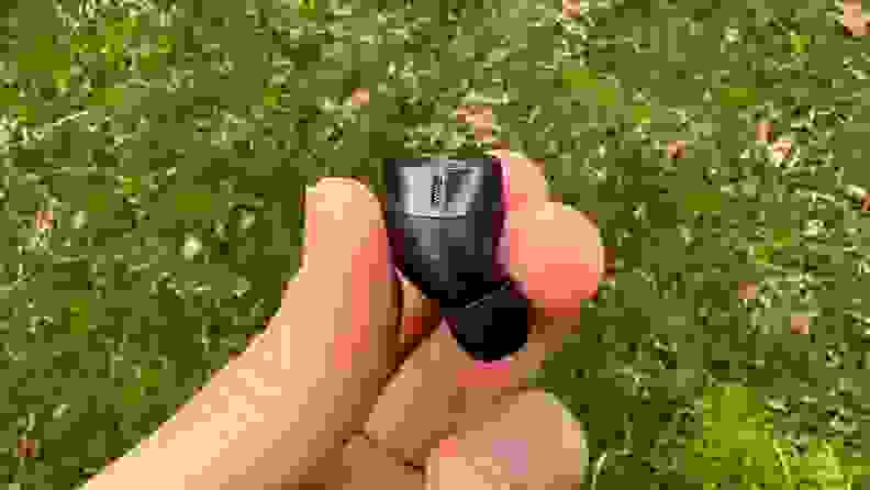 The gunsmoke colored wireless earbud sits in between two fingers, with a vent at the base and grass in the background.