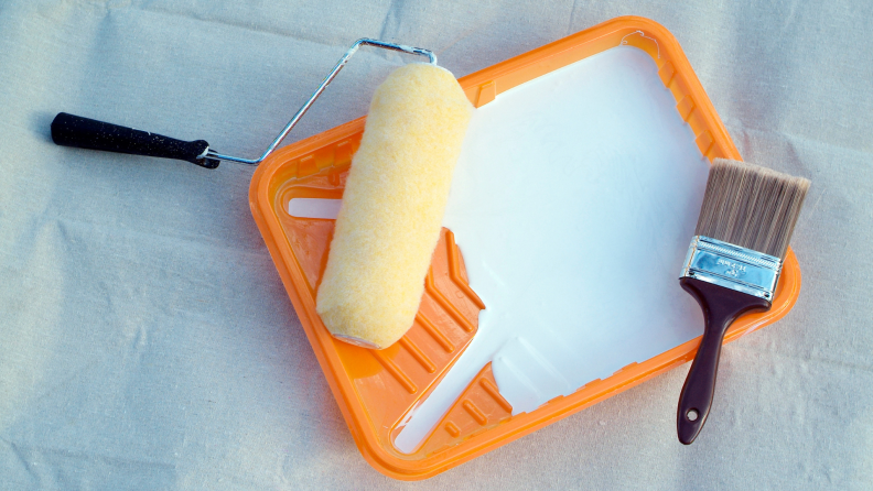 A roller and paint brush sit atop a painter's tray full of white paint.